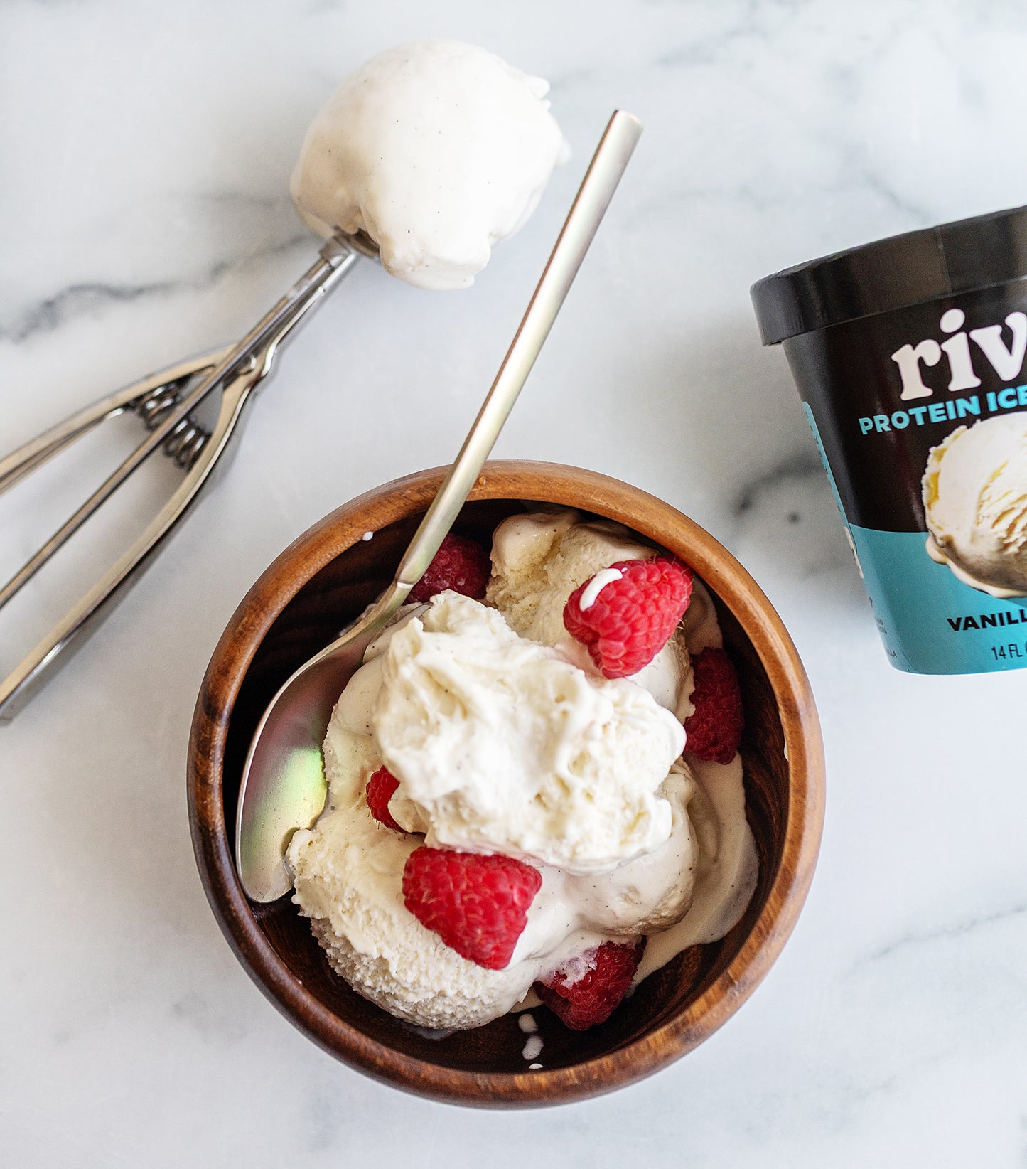 Bowl of RIvo vanilla bean ice cream with raspberries and a container of Rival Protein Ice Cream on a marble surface.