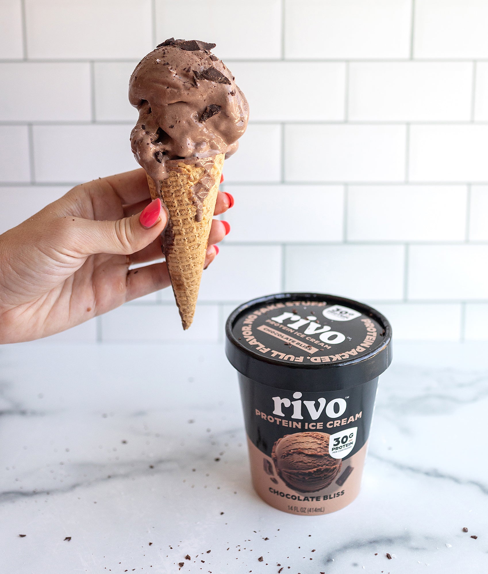 Hand holding a chocolate ice cream cone next to a container of Rivo Chocolate Bliss Protein Ice Cream on a marble surface.